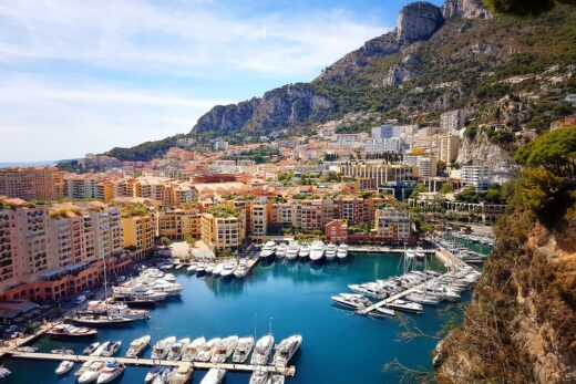 Vue panoramique d'un port de plaisance à Monaco, avec de nombreux yachts amarrés dans une eau bleue scintillante. En arrière-plan, des immeubles colorés s'étendent jusqu'aux montagnes verdoyantes sous un ciel bleu clair.